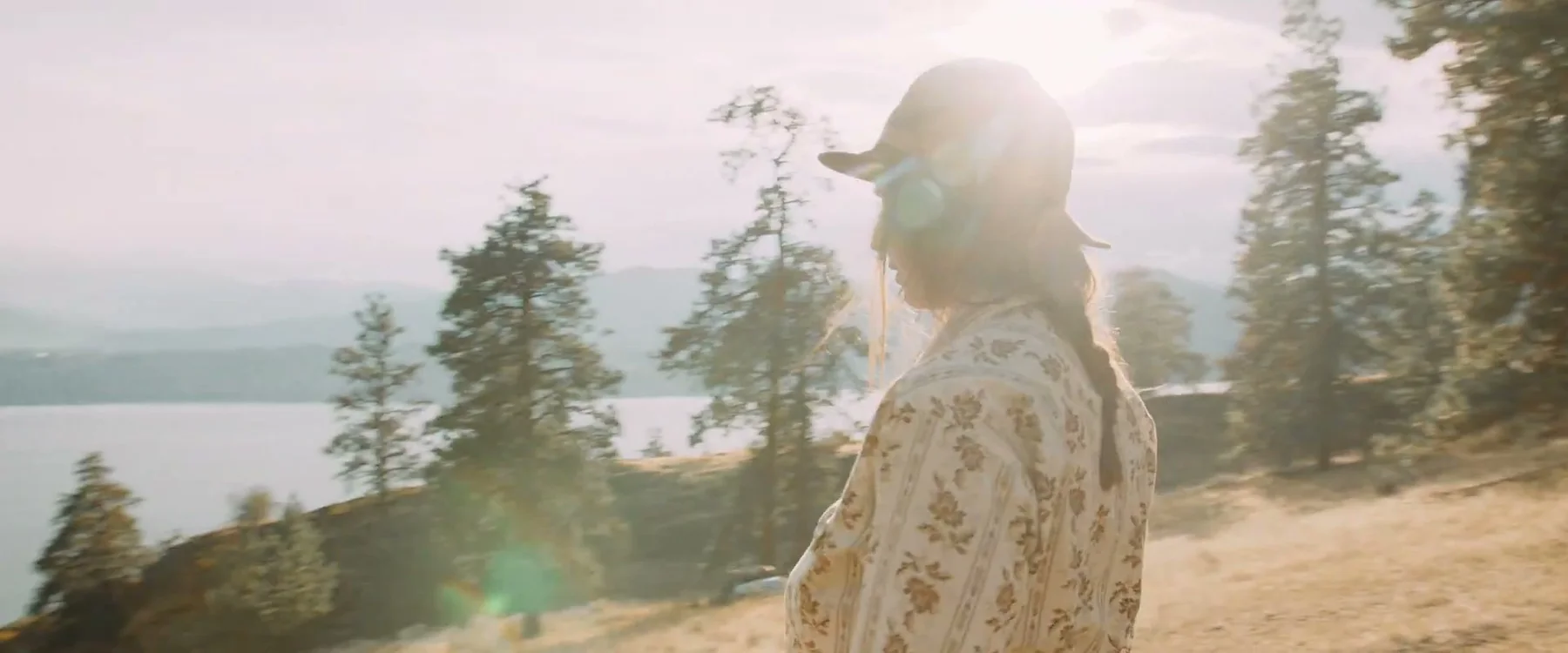 A woman in a wide-brimmed hat walks along a sunlit hillside above a lake.