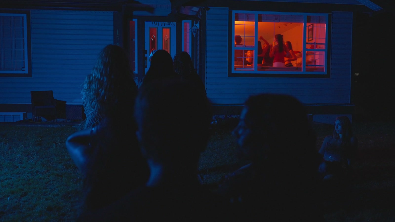 People stand outside a blue-lit house while a party glows orange-red through the window in Jamie.