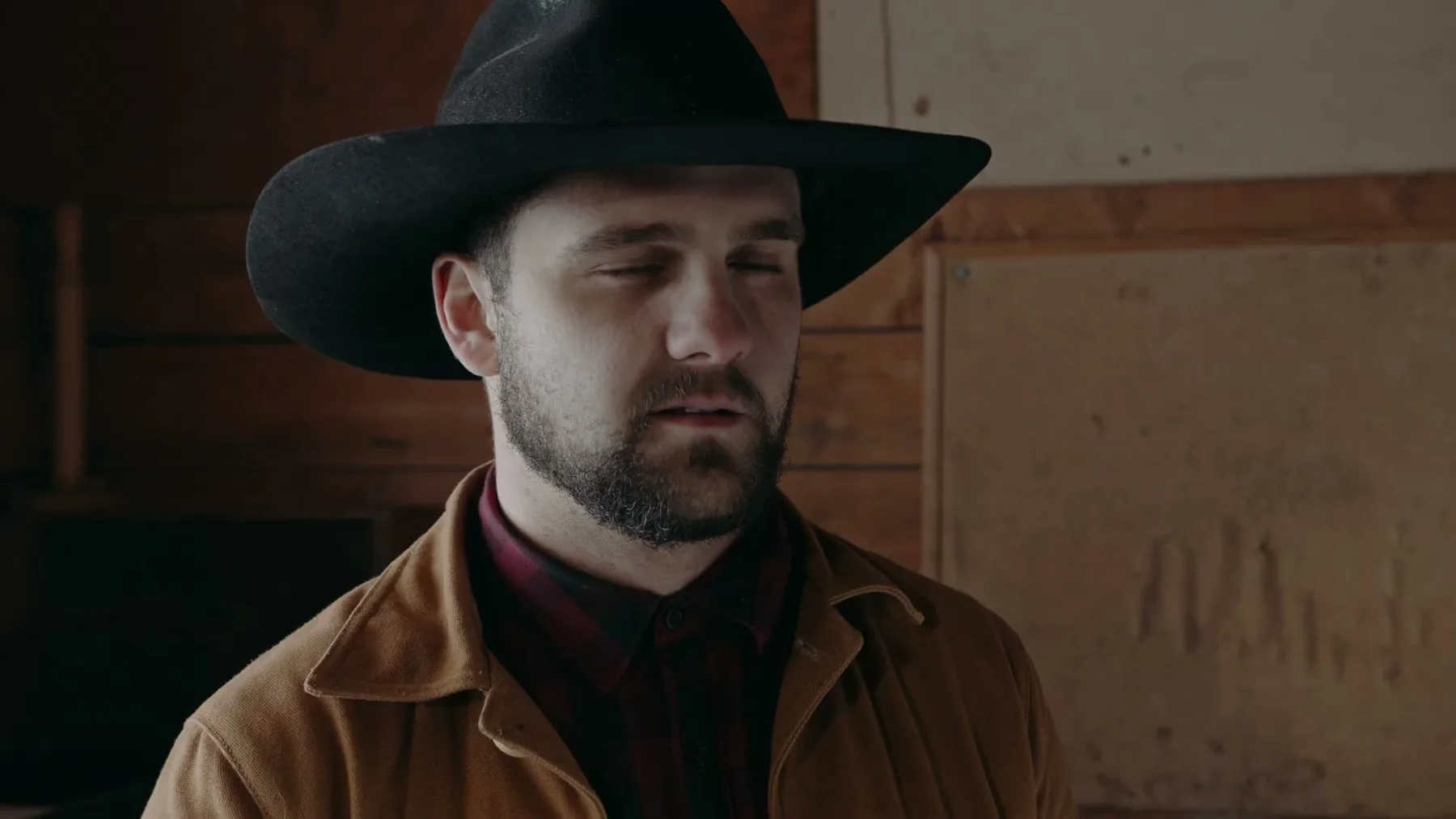 A younger man in a black cowboy hat stands against a wooden wall.
