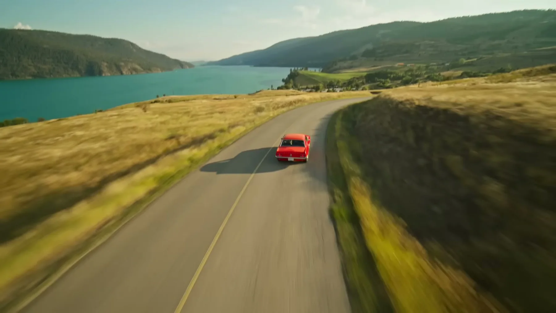 A red convertible travels along a road beside a lake and dry hills.
