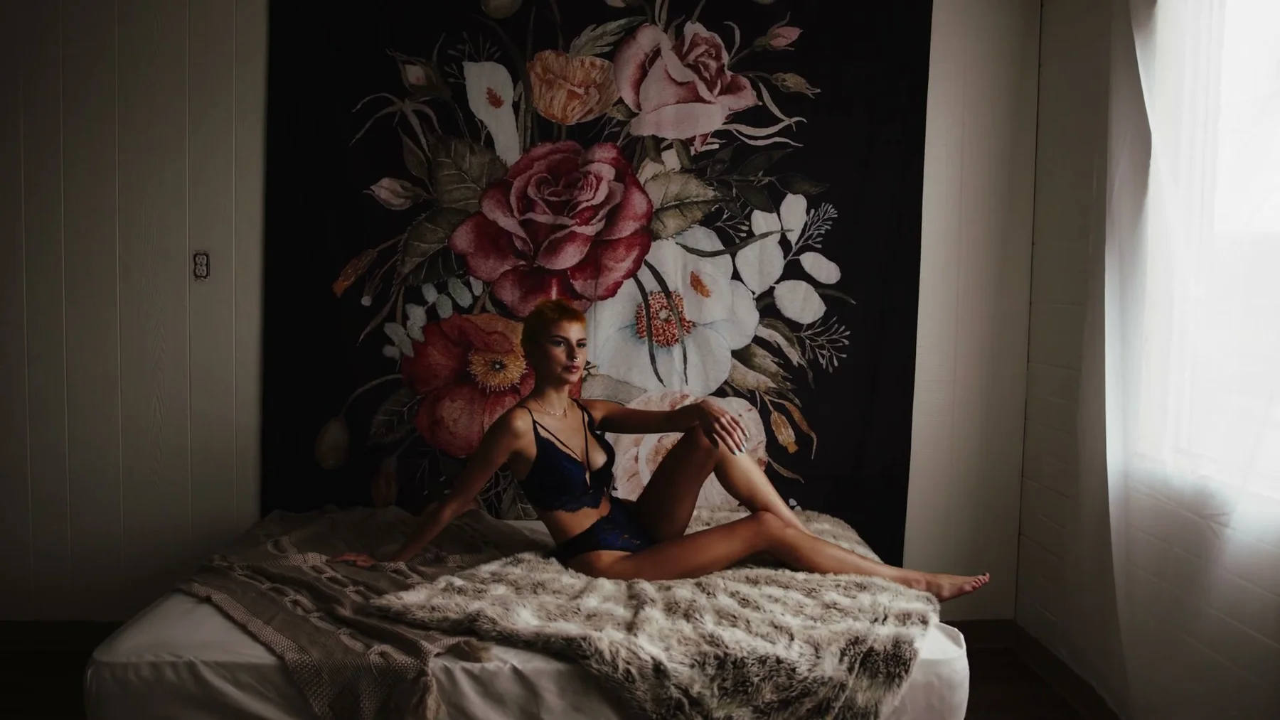 A model sits on a bed in blue lingerie beside floral wallpaper.