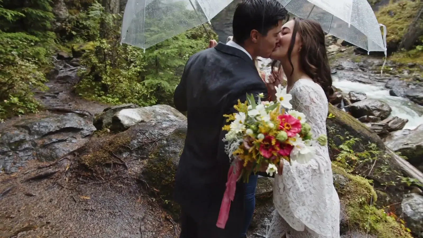 A couple kisses beneath clear umbrellas beside a waterfall.