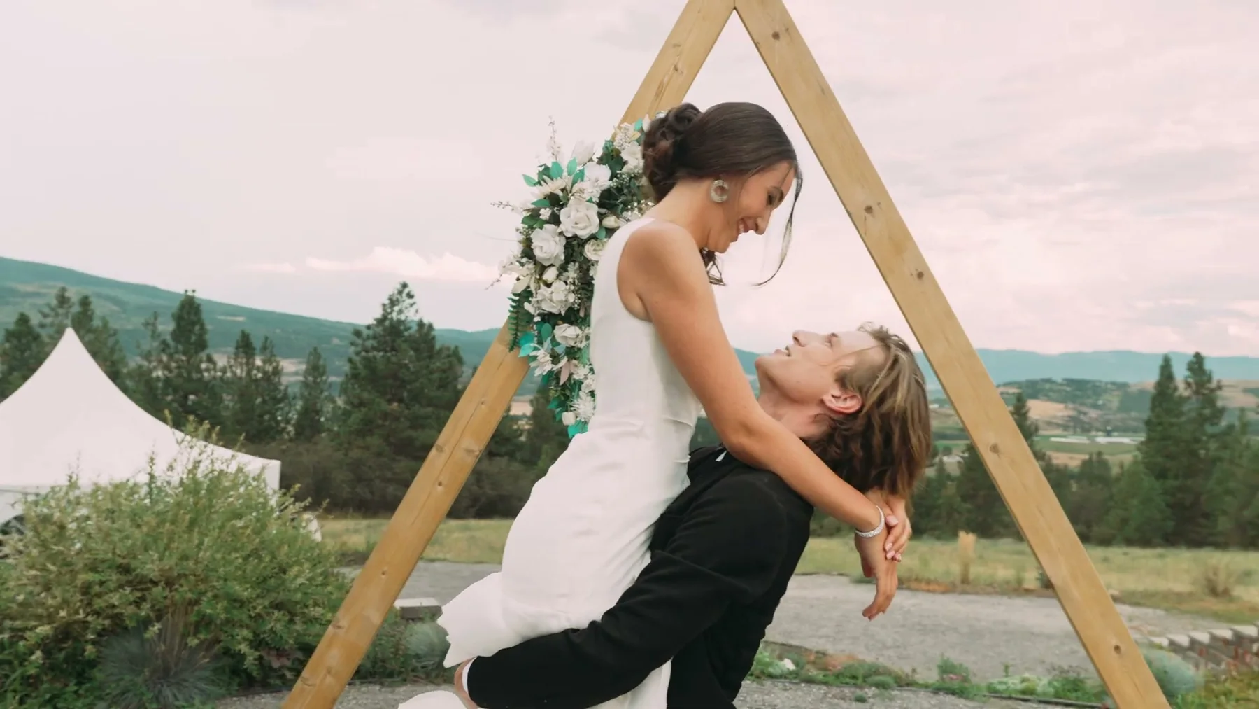 A groom dips the bride beneath a wooden wedding arch.