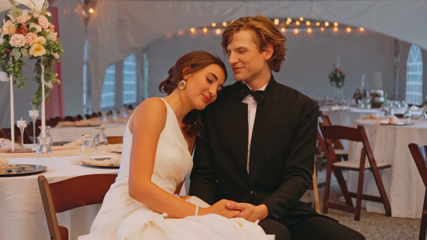 A bride rests her head on the groom's shoulder inside the reception tent.