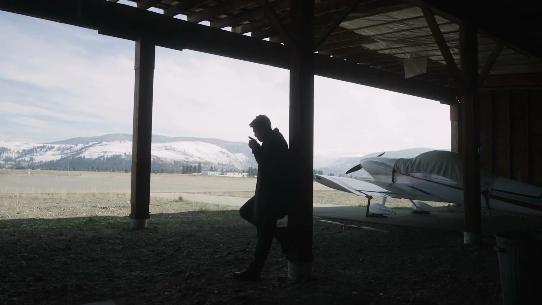 A man leans against a post inside an open aircraft hangar.